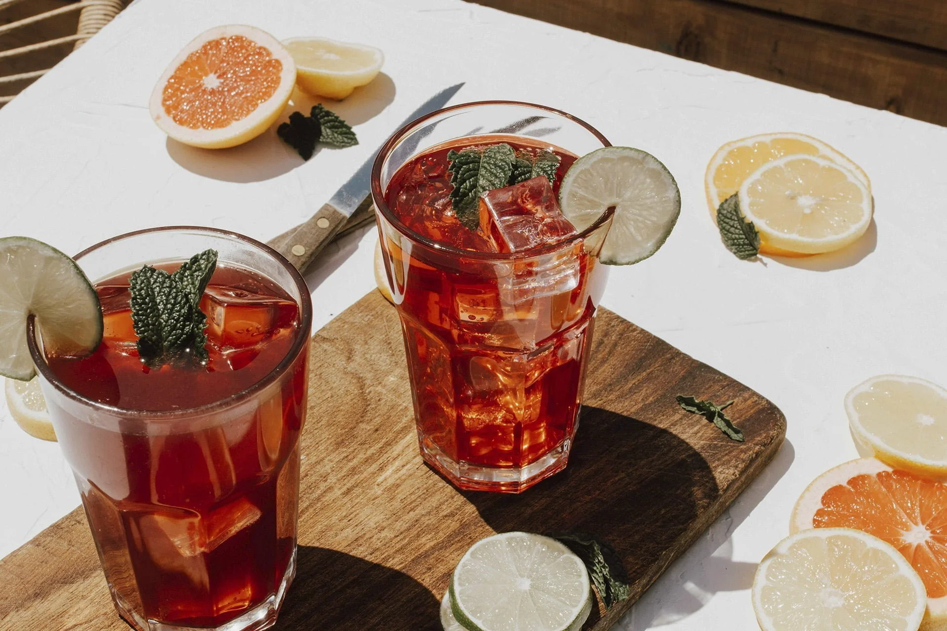 Two highball glasses filled with brown spirits cocktails with ice, slices of orange and lime, and a mint leaf on top.